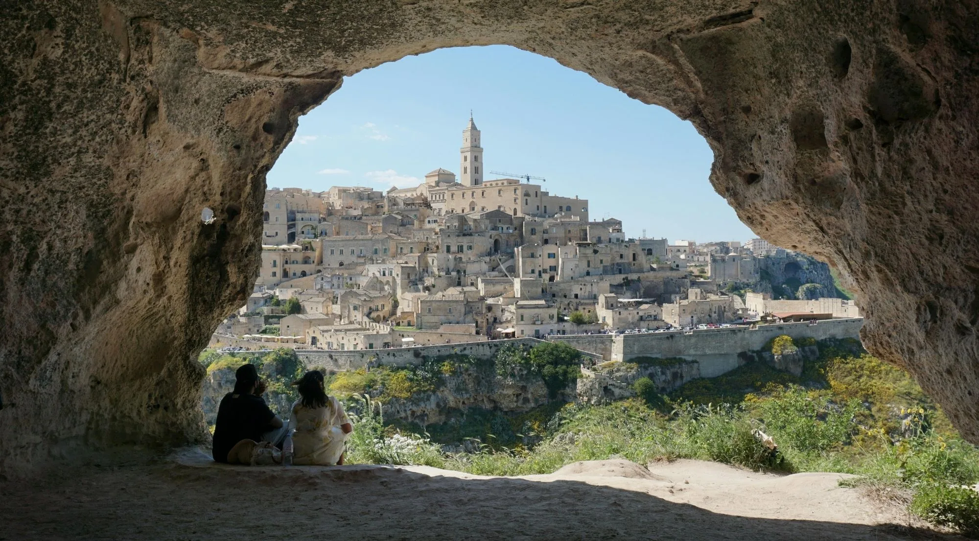 Two visitors sitting inside a cave on the Murgia plateau, looking across the gorge to Matera framed by the rock