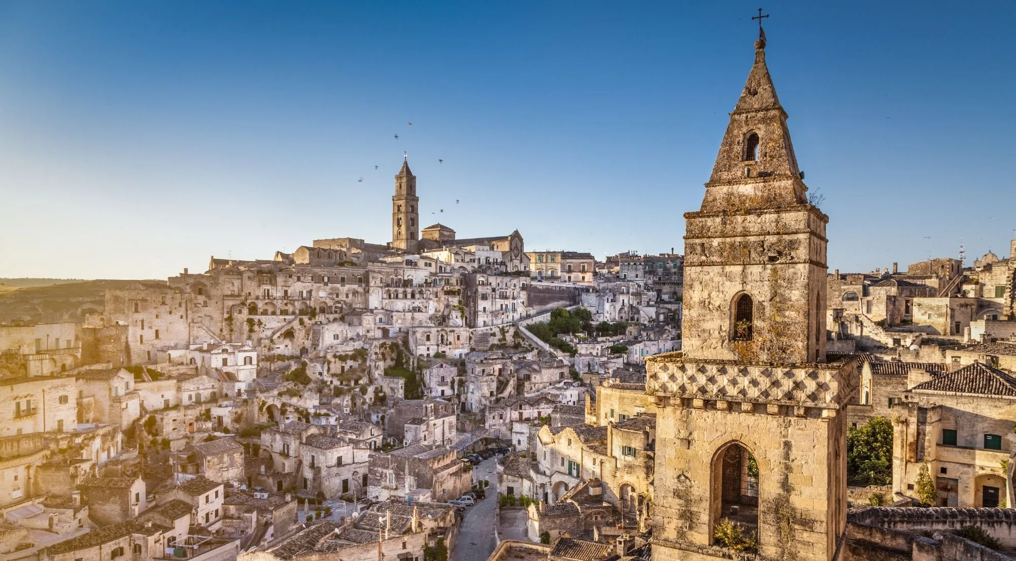 A stone bell tower in the foreground with the rooftops of the Sassi and the cathedral beyond under a blue sky