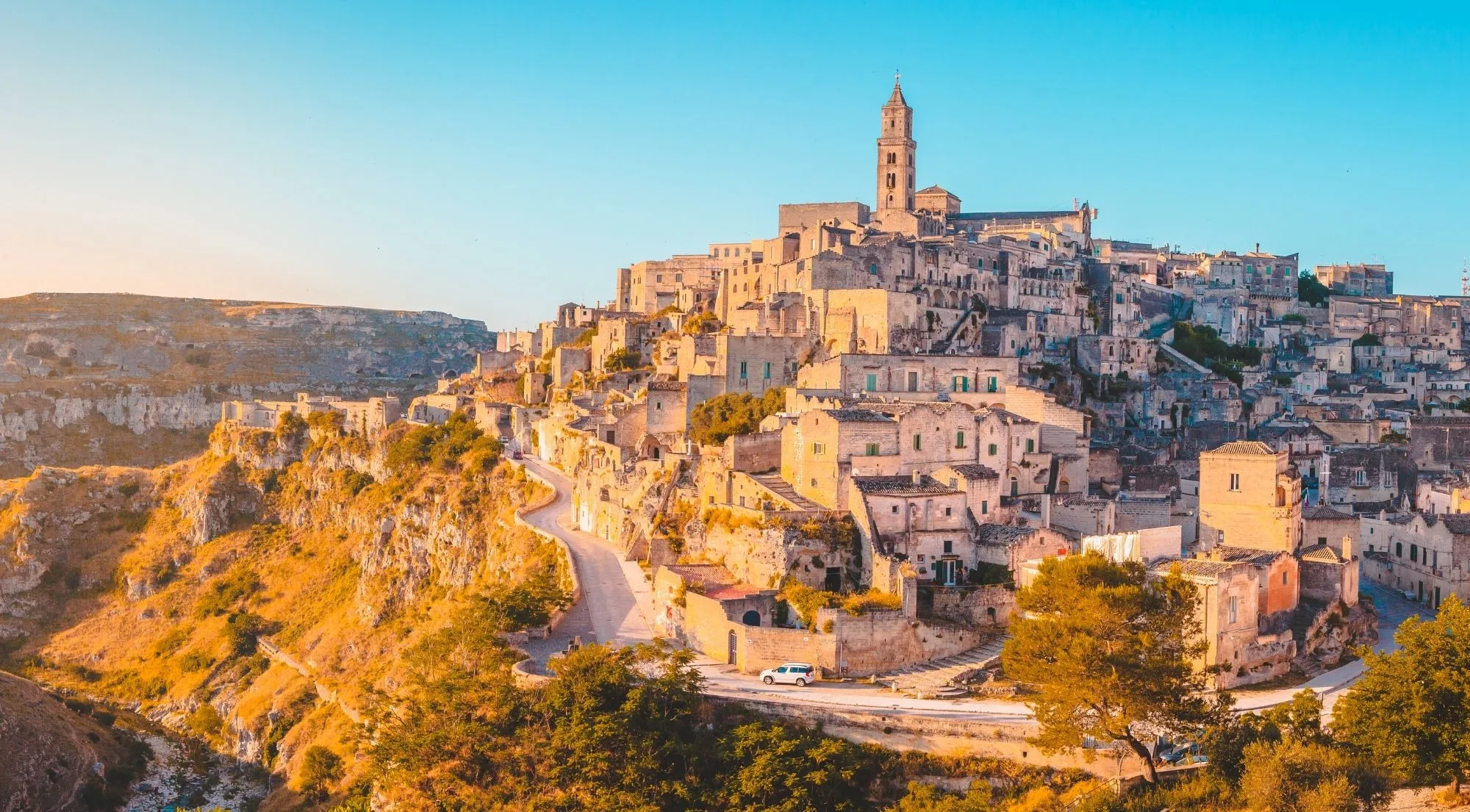 The Sassi of Matera rising above the Gravina gorge in warm evening light, with the cathedral bell tower at the top