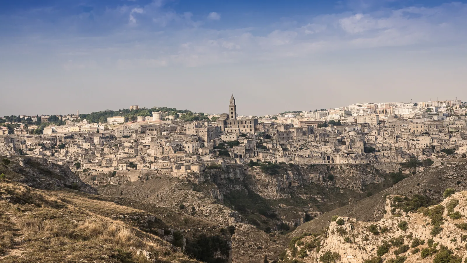 A wide view of Matera at dusk across the gorge, with the town and bell tower along the horizon.