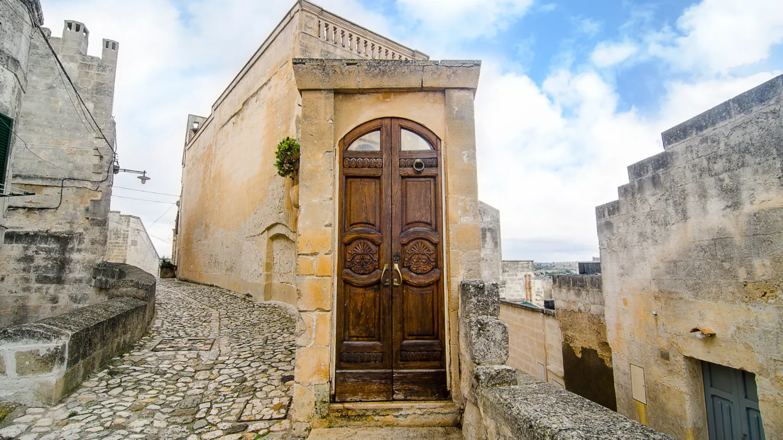 A carved wooden double door in a pale stone wall at the end of a cobbled lane in the Sassi.
