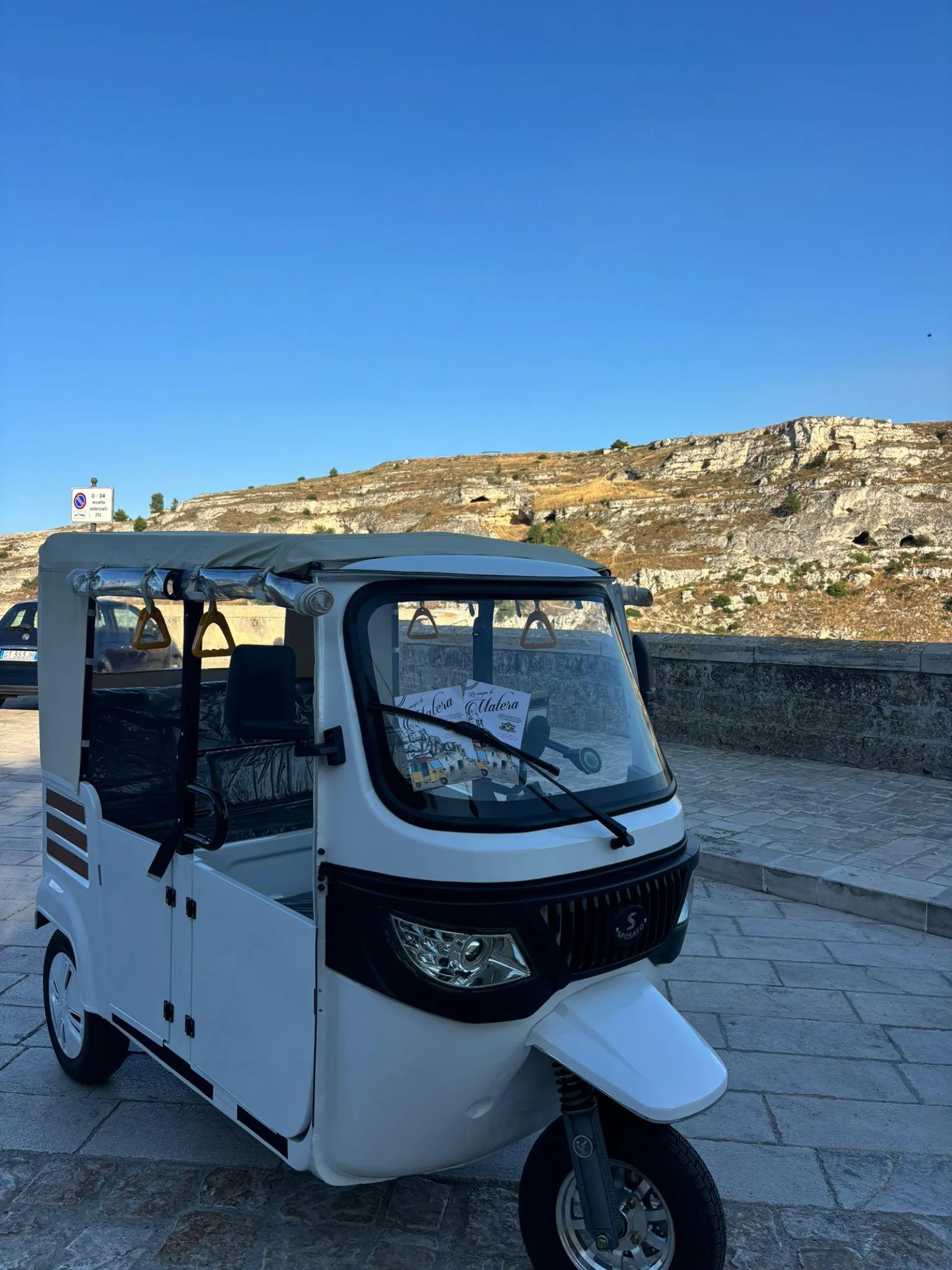 A white electric tuk-tuk parked on a paved lookout with the rocky Murgia hillside behind it.