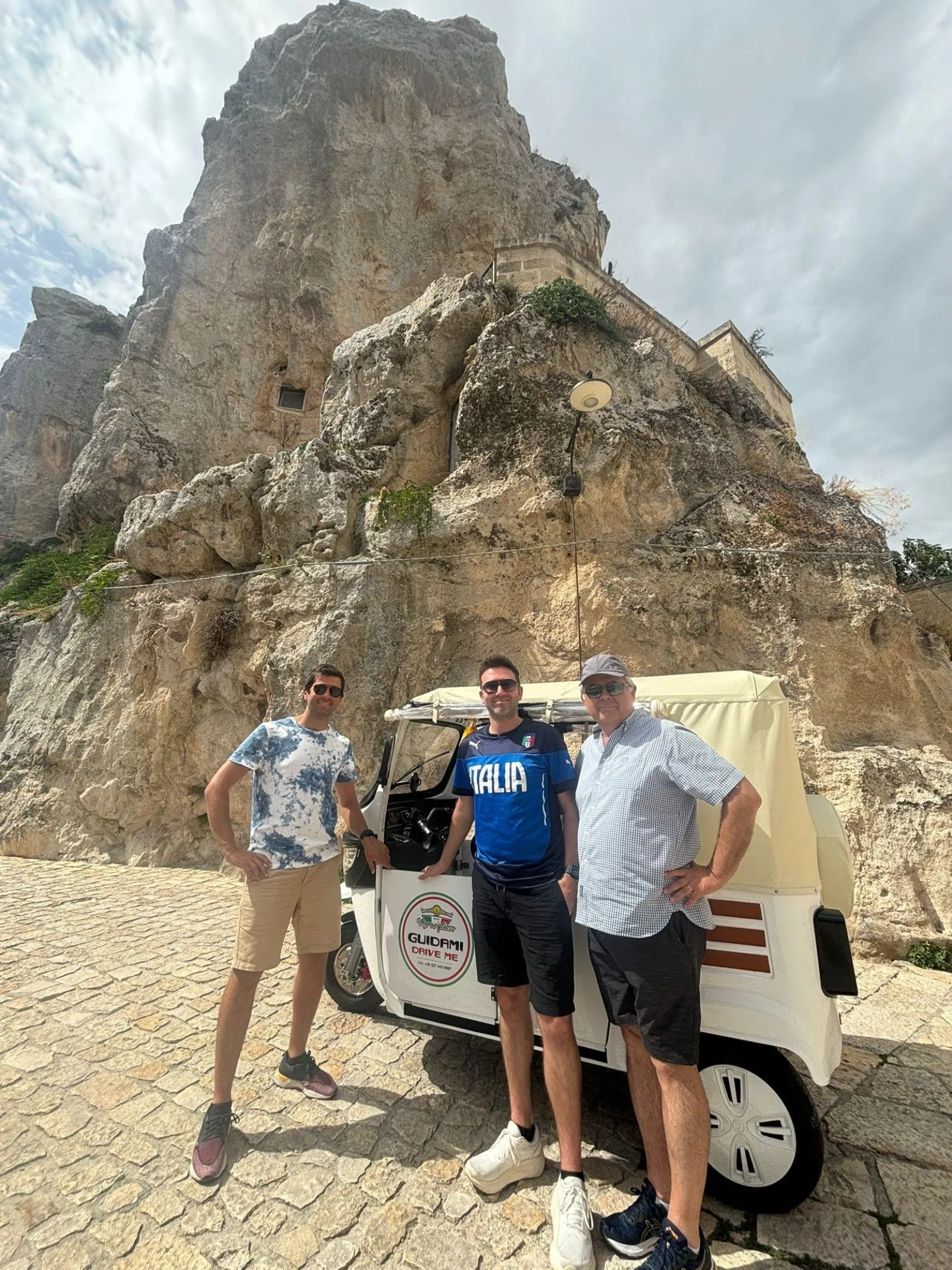 Three men stand beside a cream and white tuk-tuk beneath a tall limestone crag in Matera.