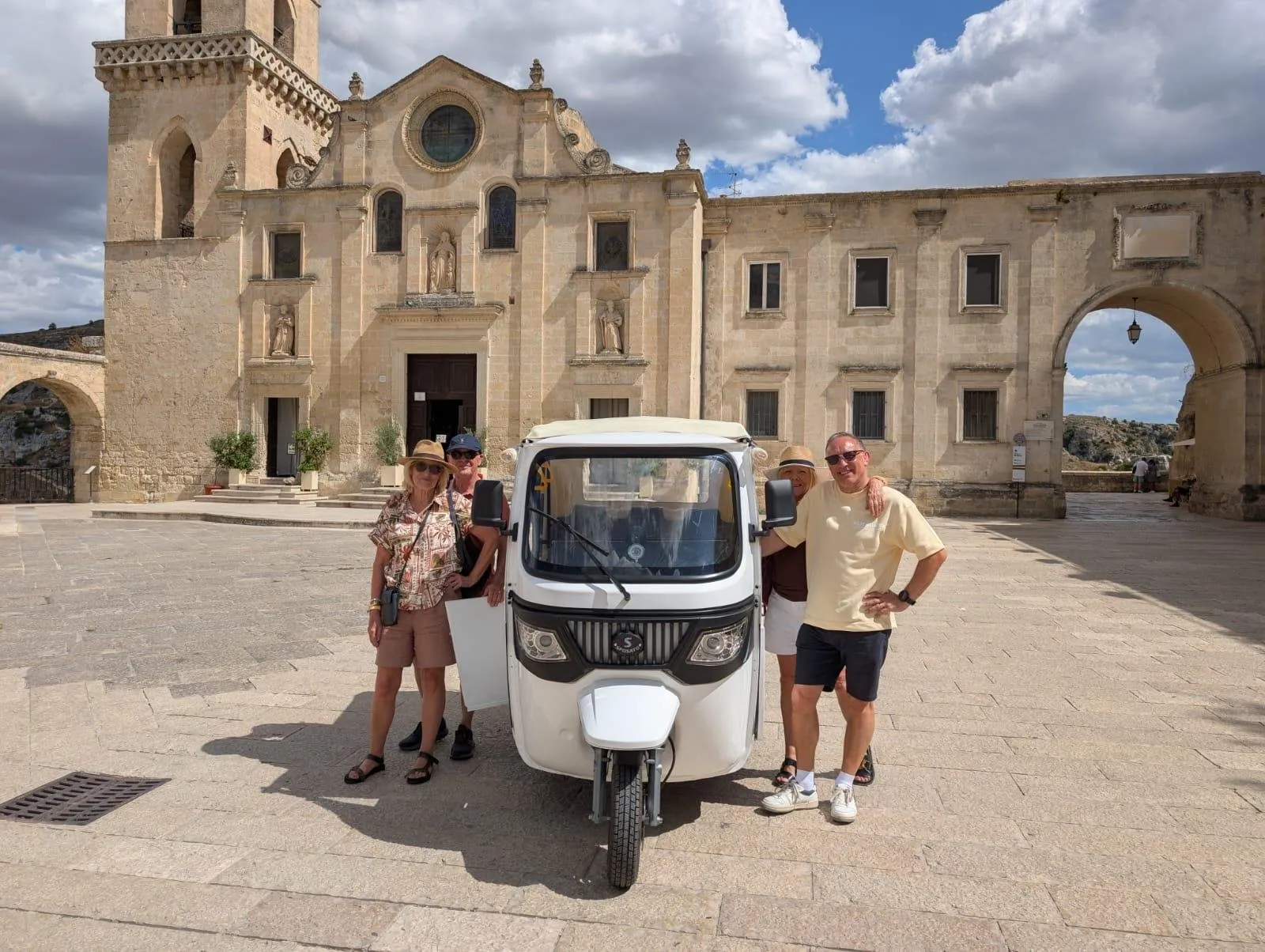 A white tuk-tuk and four visitors stand in front of a pale stone church facade with a rose window.