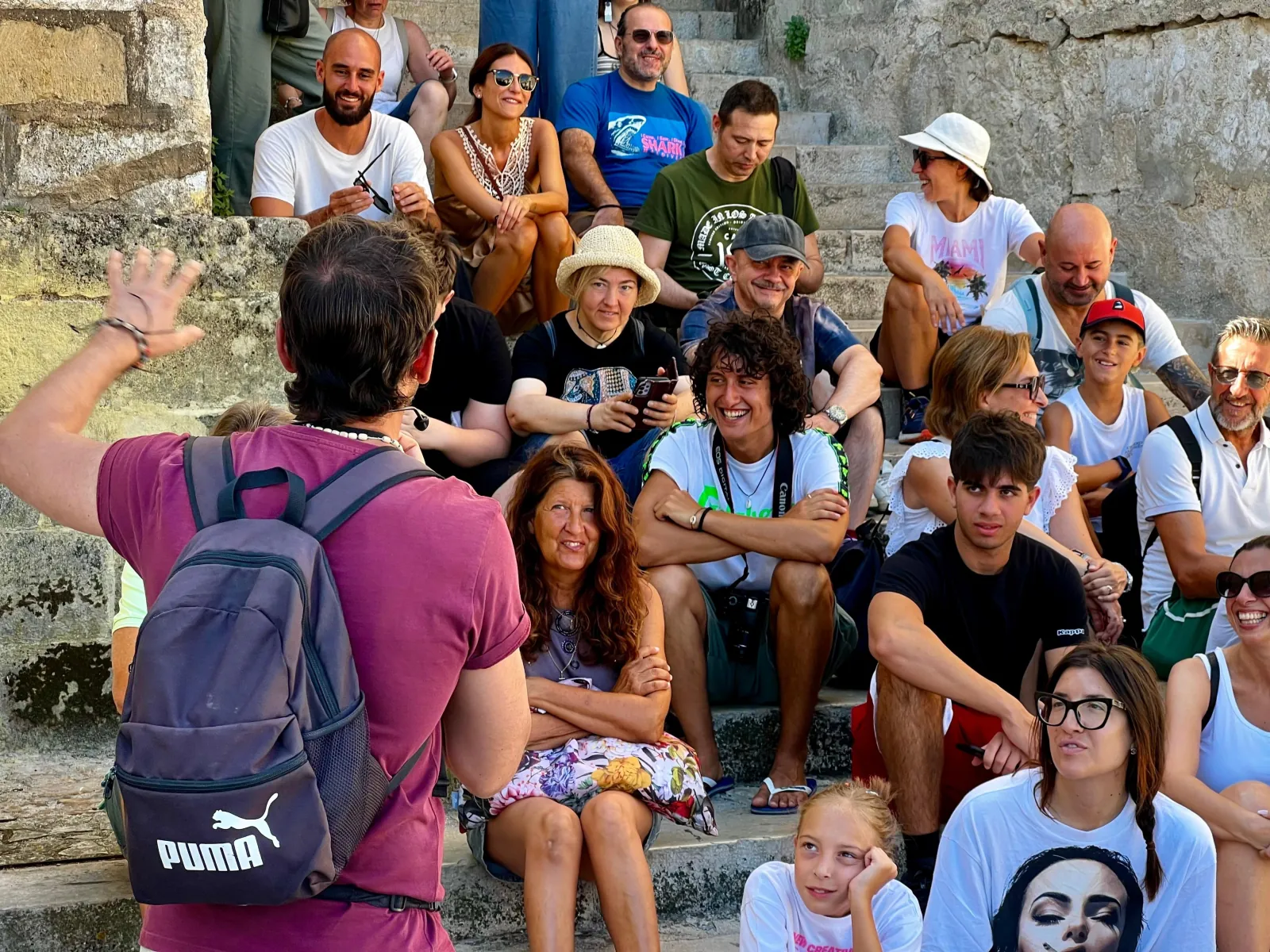 A guide in a maroon shirt addresses a large group seated on stone steps in a Matera alley.