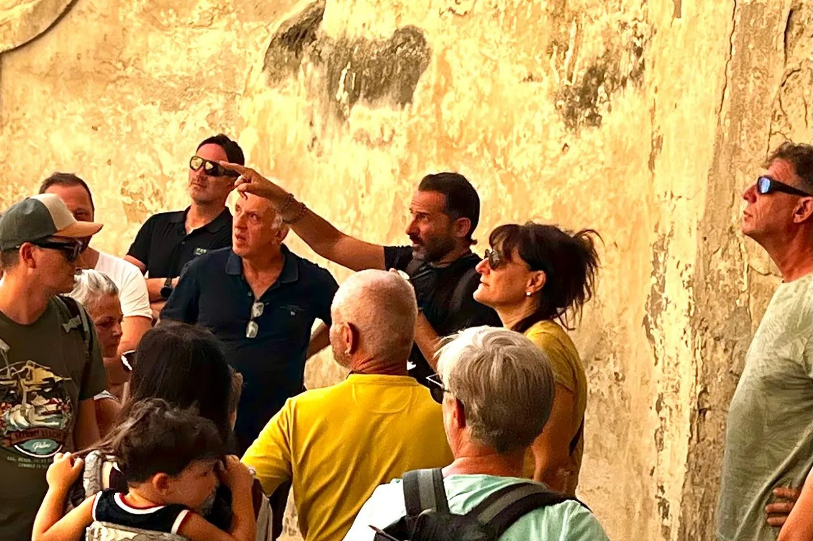 Tour participants crowd around their guide, who points upward, beside a weathered golden-yellow wall in Matera.