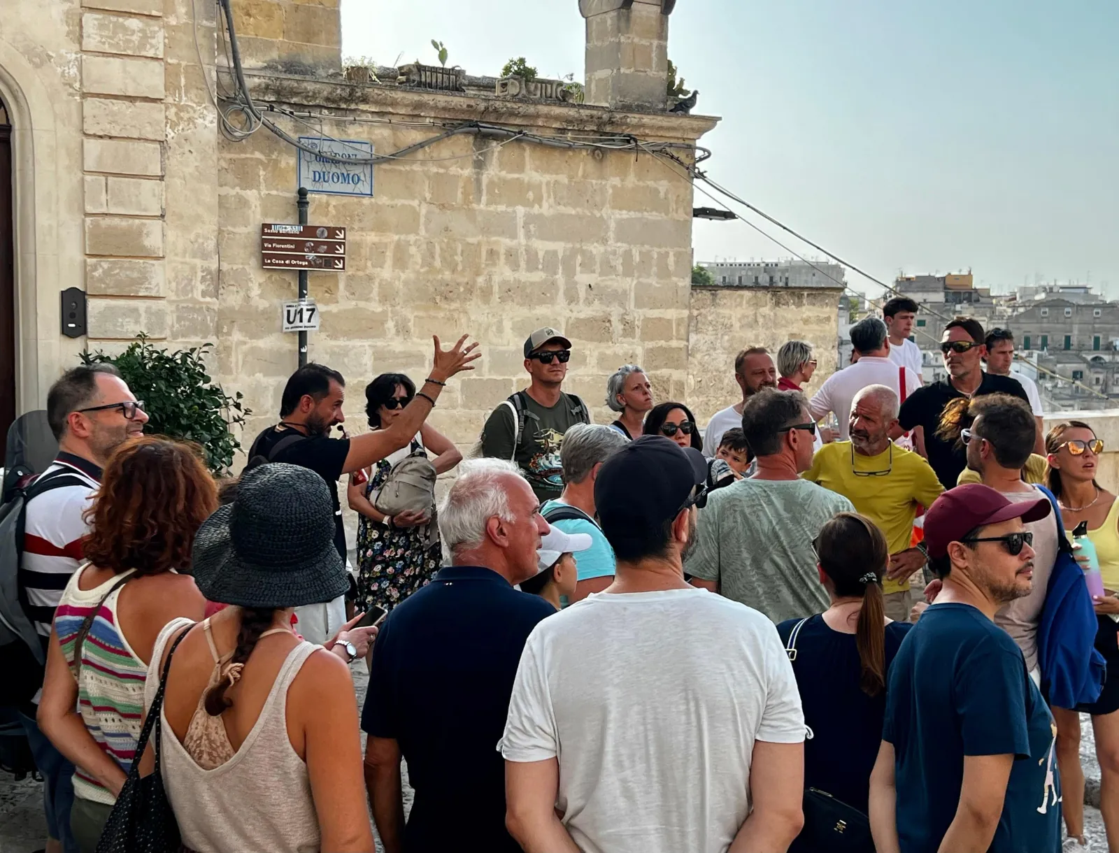 A large group listens to a guide who points upward on a street corner in Matera's old town.