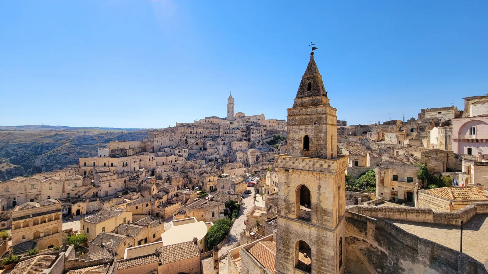 A church bell tower in the foreground over the pale stone houses of the Sassi di Matera under blue sky.