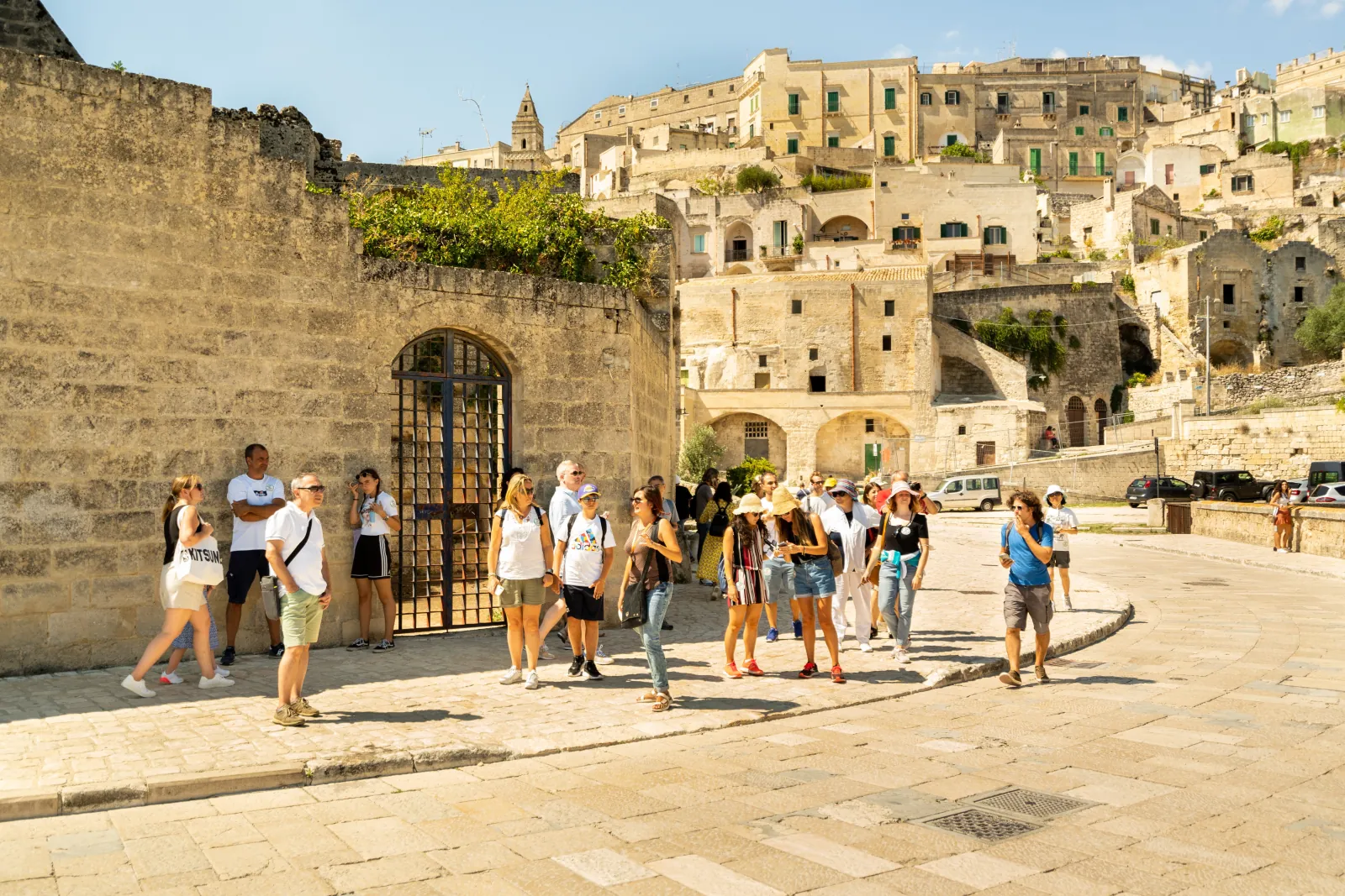 A tour group gathers on a sunlit stone square below the tiered stone houses of the Sassi in Matera.