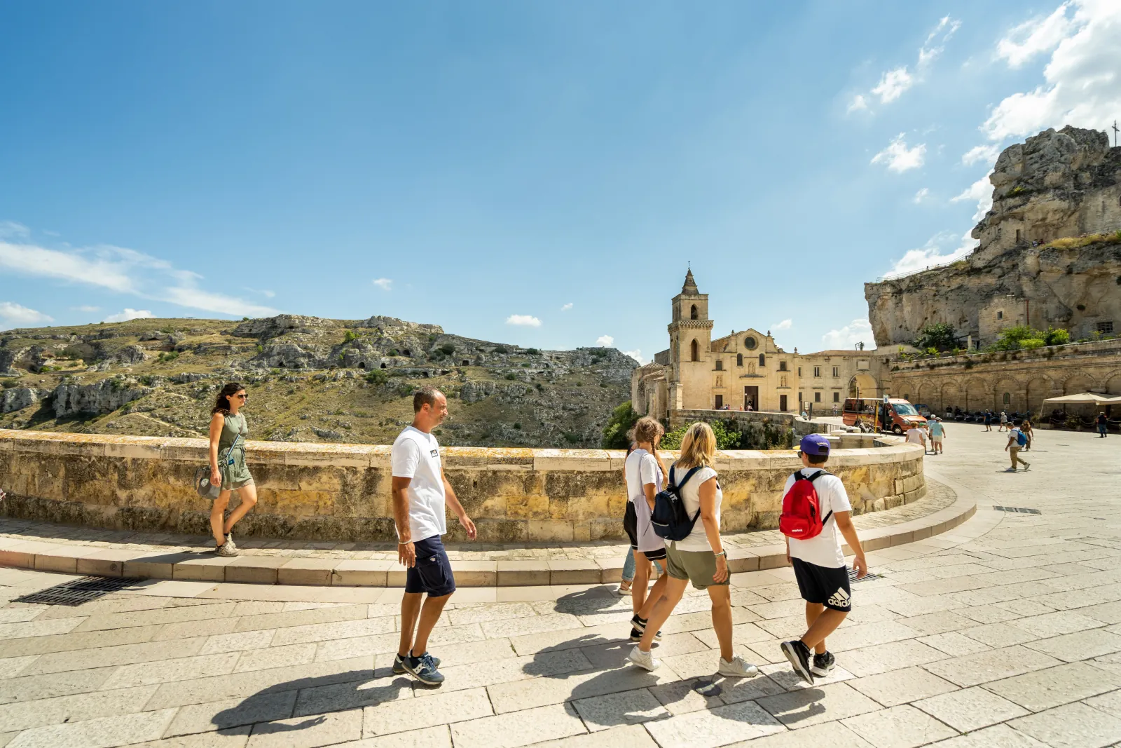 Visitors walk across a paved terrace toward a small church with a bell tower, with the rocky gorge behind.