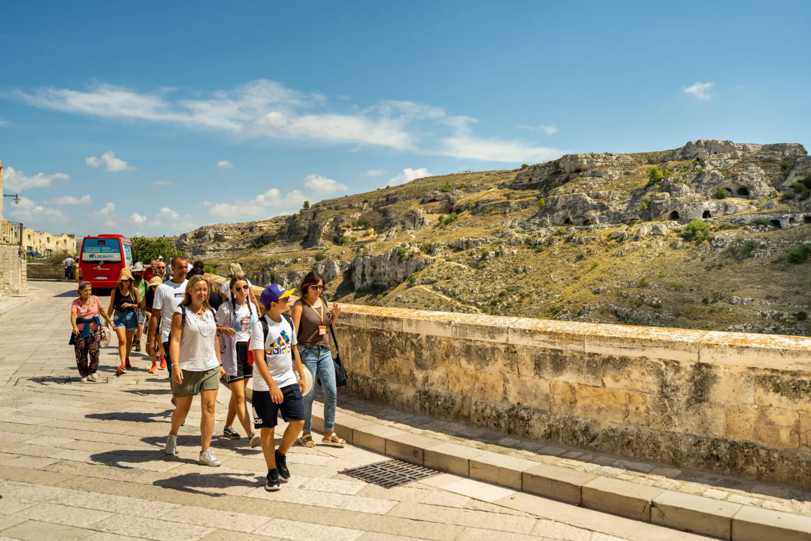 A tour group walking along a stone road above the gorge, with cave-dotted Murgia slopes opposite