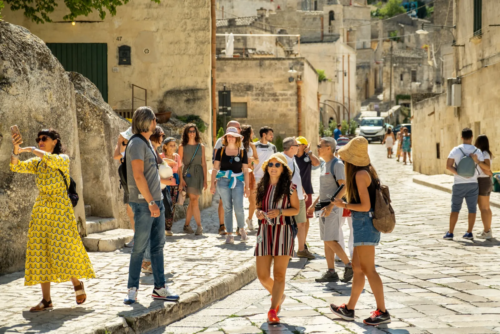 Visitors in summer clothes walk and take photos along a sunlit cobbled lane of pale stone houses in the Sassi.