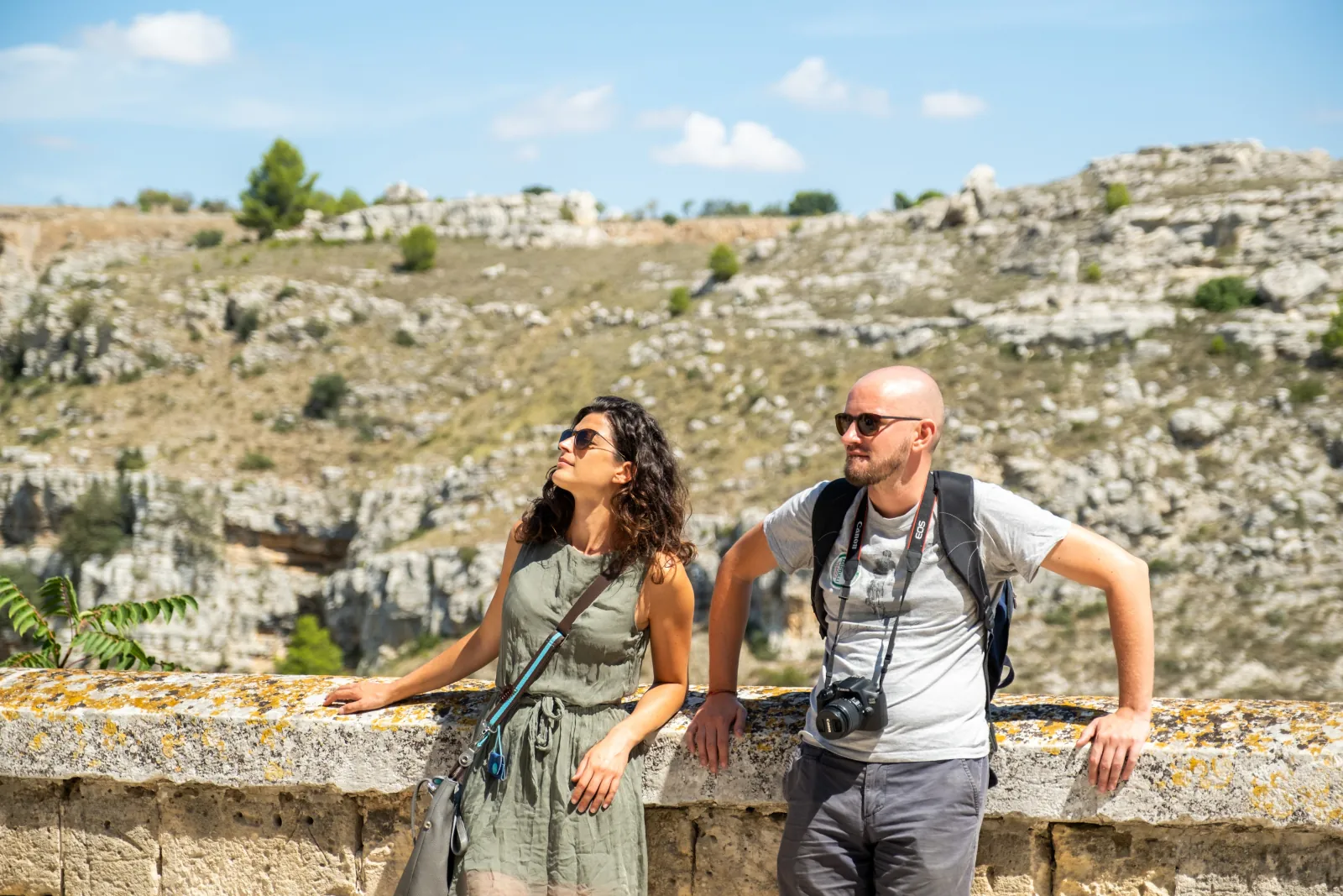A woman and a man with a camera lean on a stone wall above the rocky Murgia hillside.