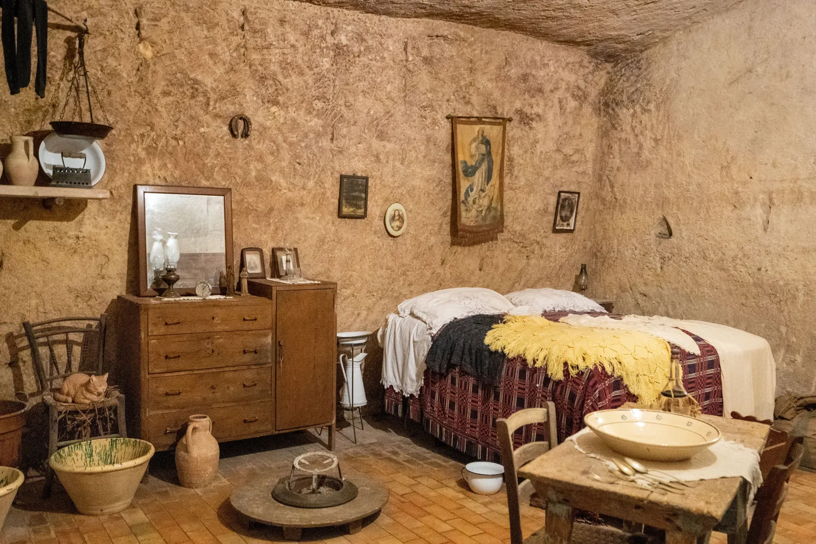 A furnished cave house with a bed, wooden chest of drawers and table under a rough rock ceiling in Matera.