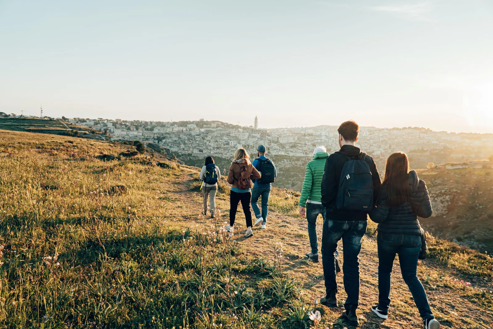 Hikers walk across a golden grassy hillside on the Murgia plateau toward Matera at sunset.