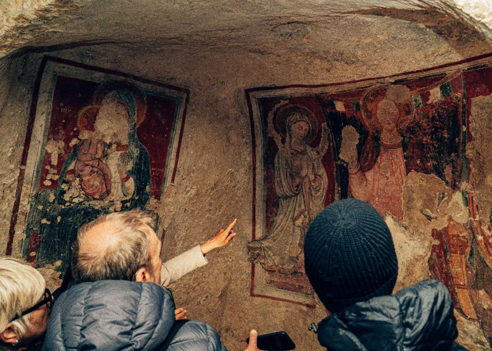 A guide points out faded red and gold frescoes on the wall of a rock-cut church to a group.