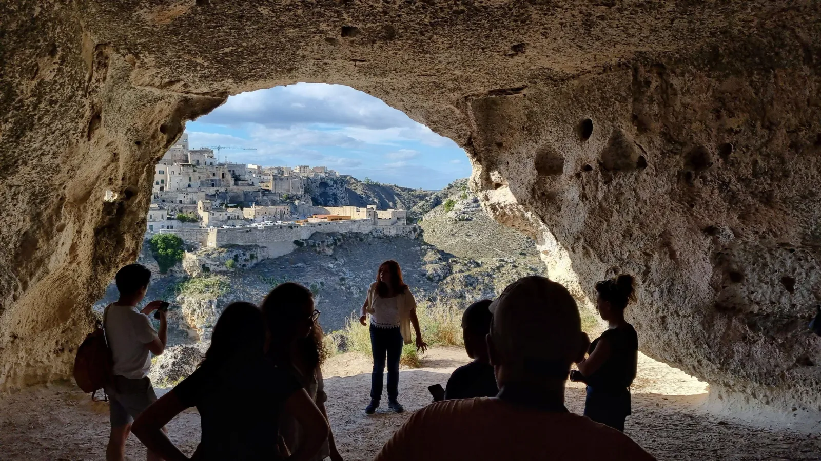Silhouetted visitors stand inside a large Murgia cave opening that frames the distant Sassi di Matera.