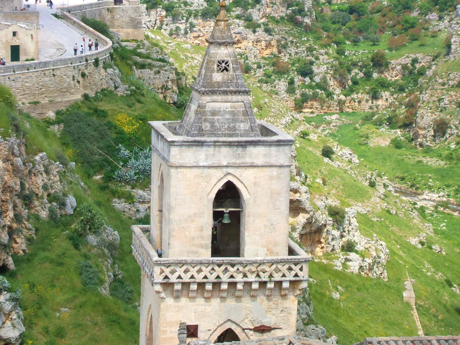 A stone bell tower with a pointed spire rises above the green slopes of the Gravina gorge near Matera.