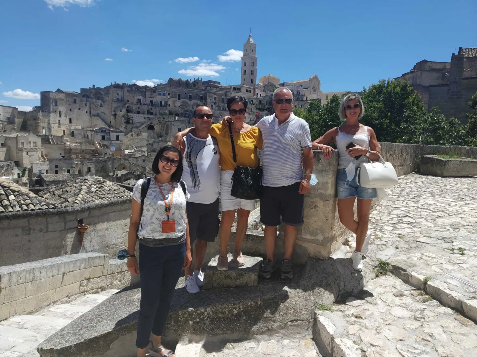Four visitors pose on a stone terrace with the Sassi and the cathedral bell tower behind them.