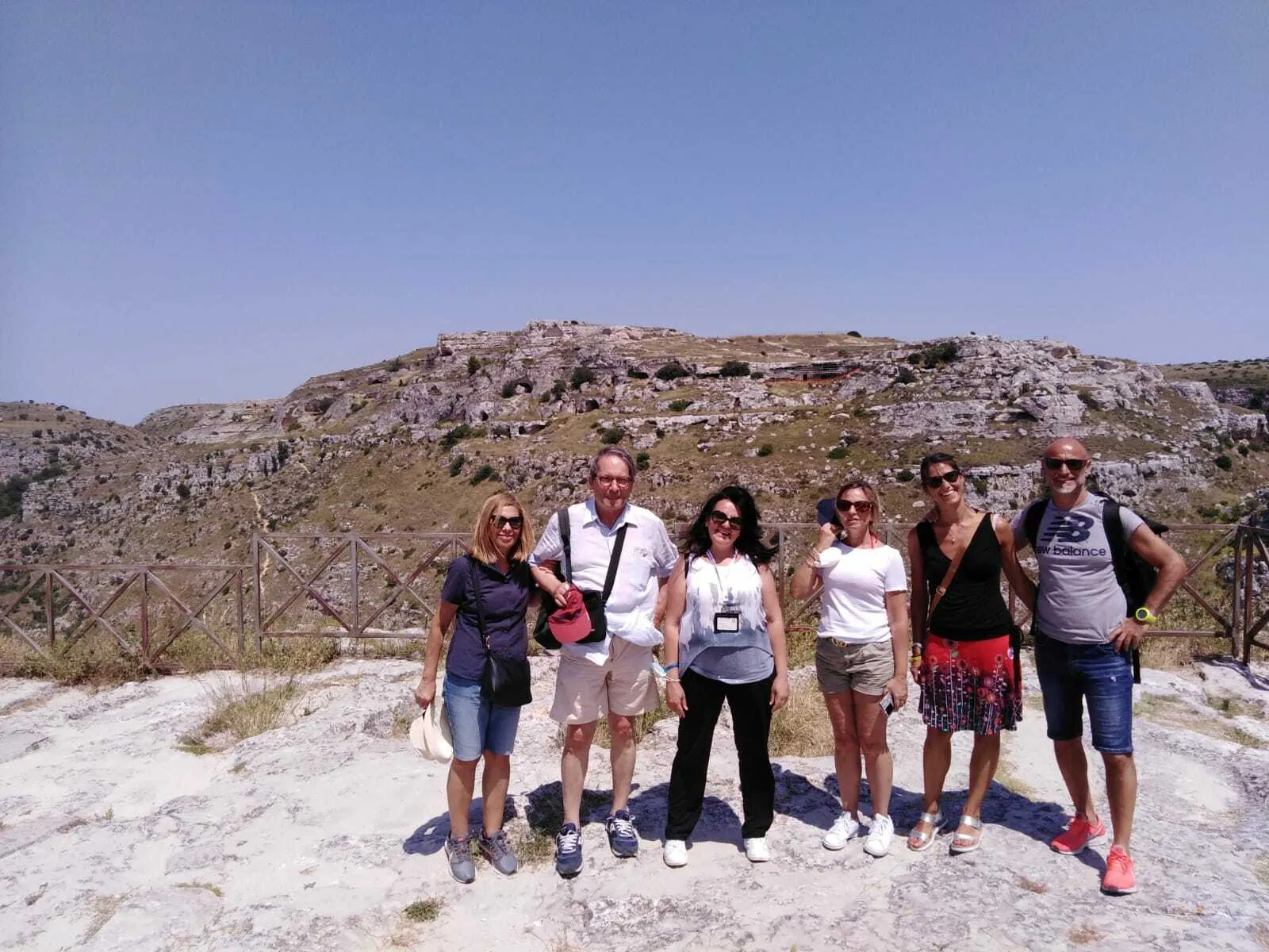 Six visitors stand on pale limestone at a viewpoint, with the Murgia hillside and its caves across the gorge.