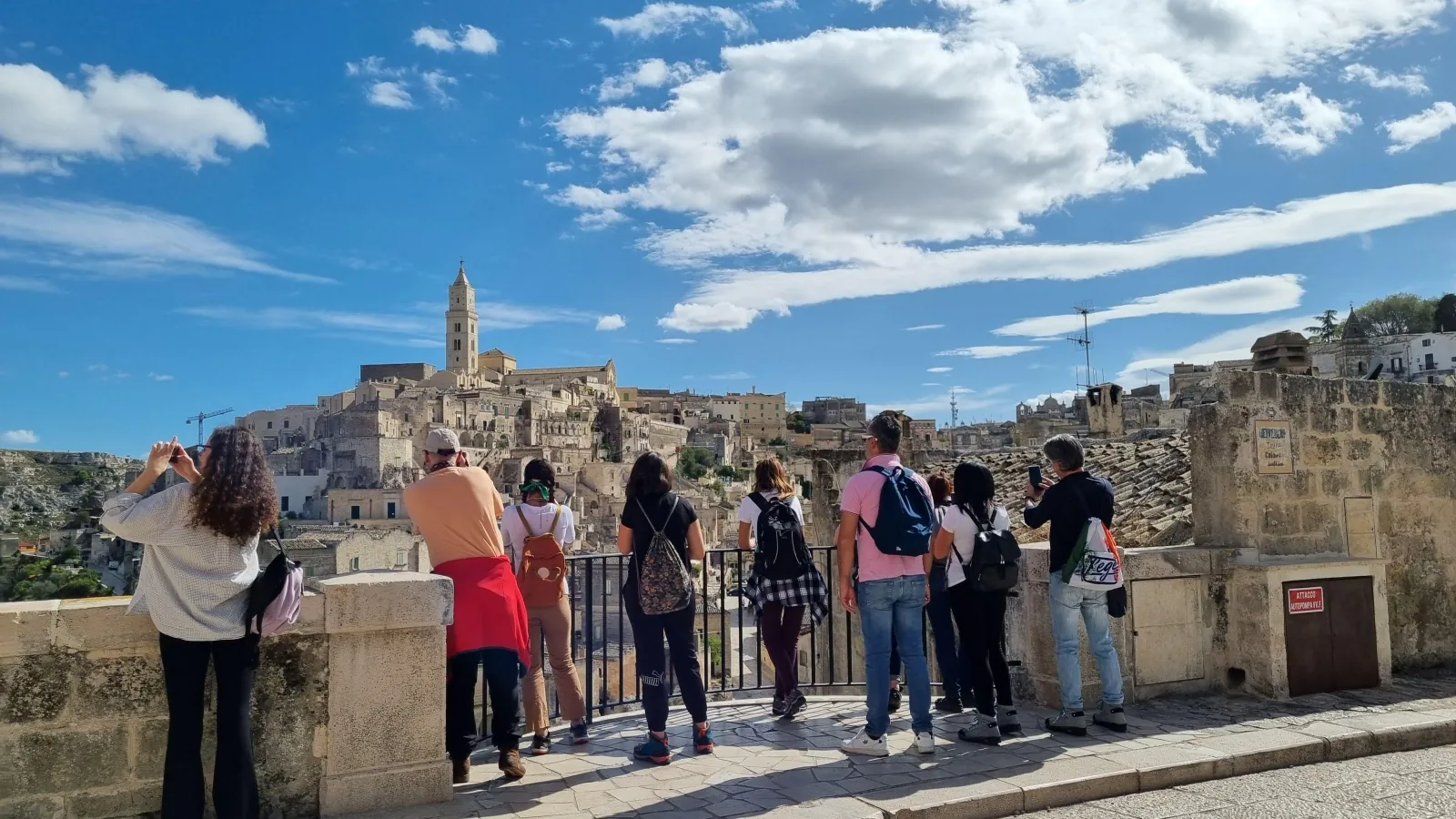Visitors photograph the Sassi and the cathedral bell tower from a stone railing on a sunny day.