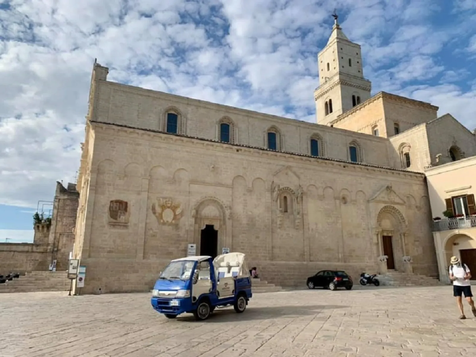 A small blue tuk-tuk crosses the square in front of Matera Cathedral and its bell tower.