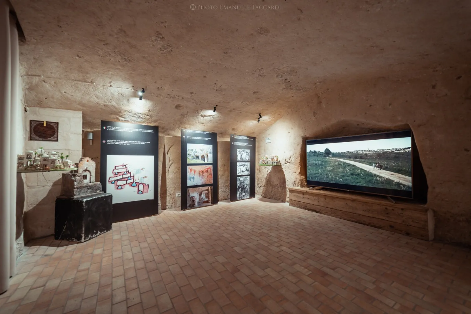 Inside a cave-house museum in Matera, display panels and a wall screen stand on a brick floor under spotlights.