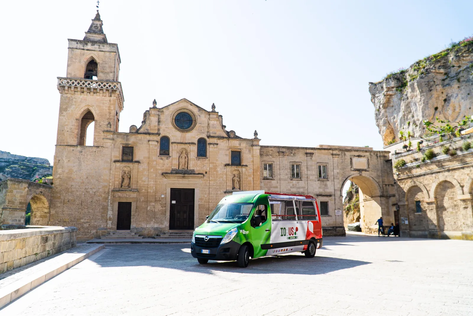 A green and red minibus stands in the square before a rock-cut church with a bell tower and arched gateway.