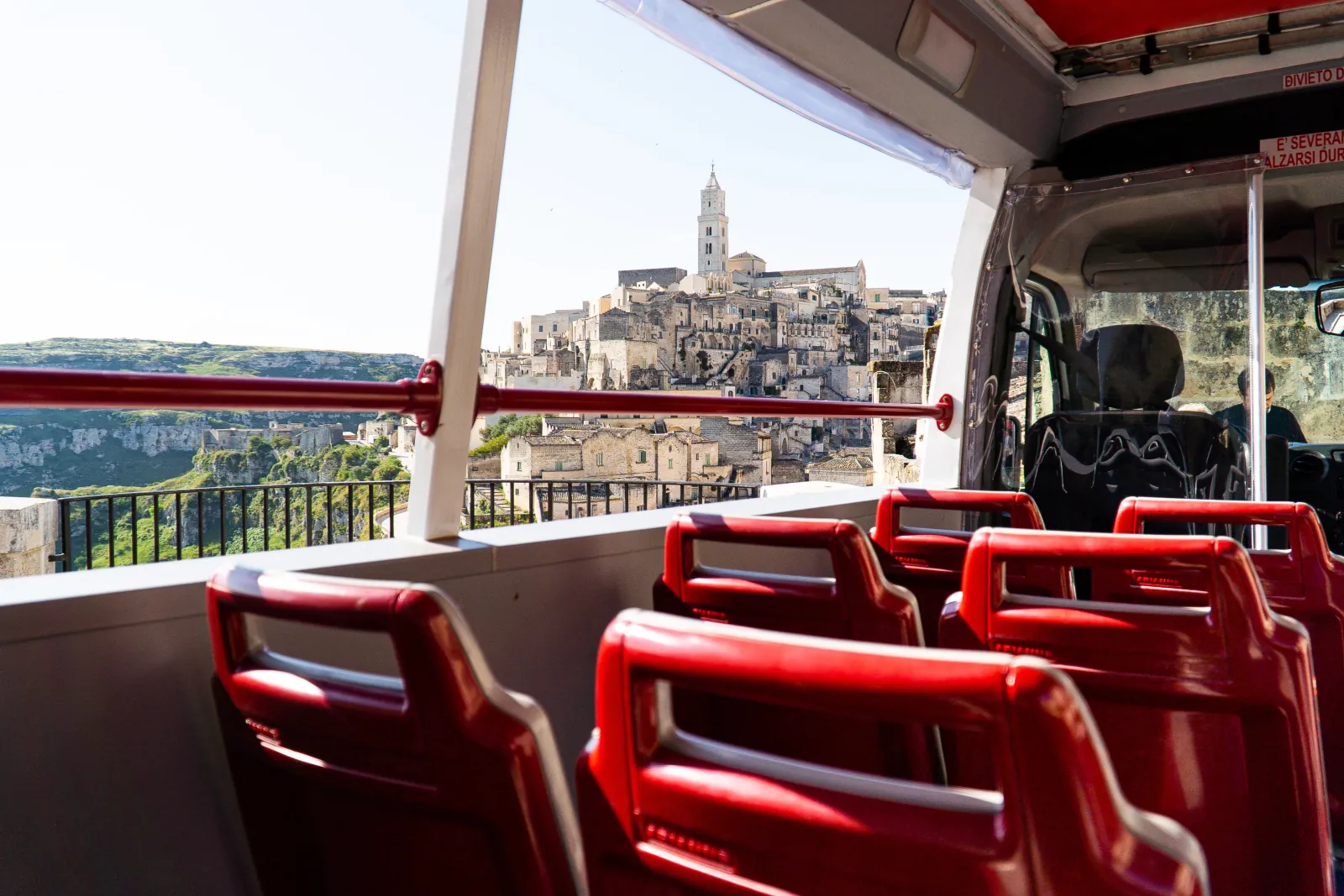 Red seats on the open upper deck of a tour bus, with the Sassi and cathedral tower beyond.