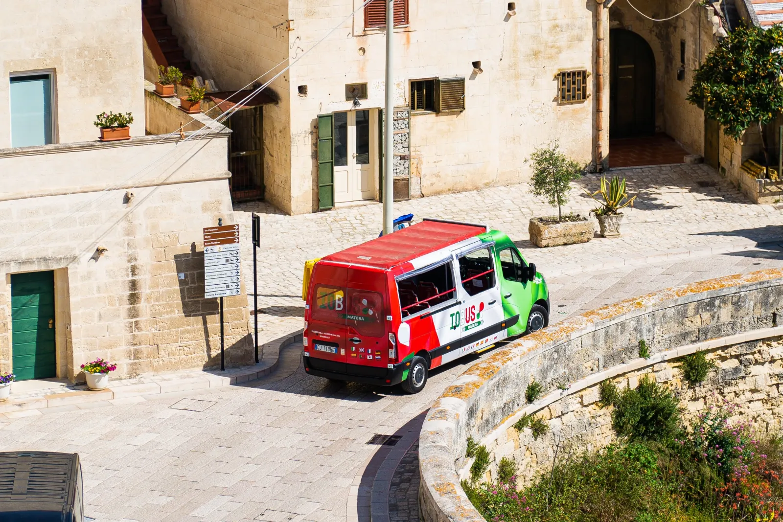 A green and red tour minibus rounds a curve between stone houses with green doors and shutters in Matera.