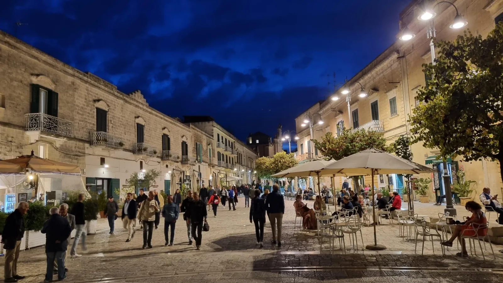 Evening on a lamp-lit Matera street, with people strolling and cafe tables under parasols beside stone buildings.