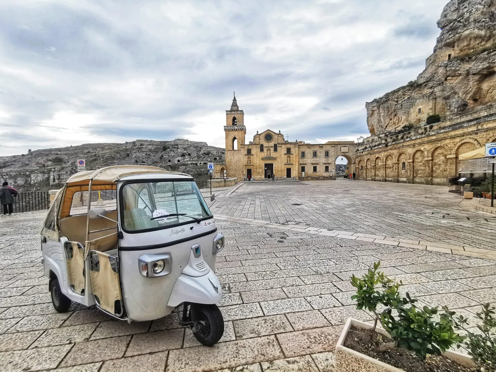 A small white three-wheeled Ape tuk-tuk parked on a stone square in front of a church with a bell tower.