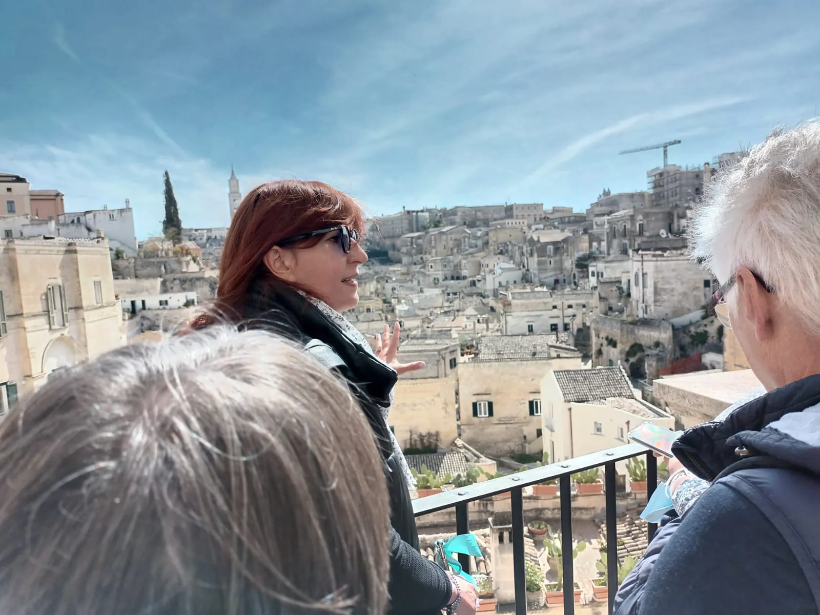 A guide in sunglasses gestures toward the Matera skyline from a railed terrace while two visitors listen.