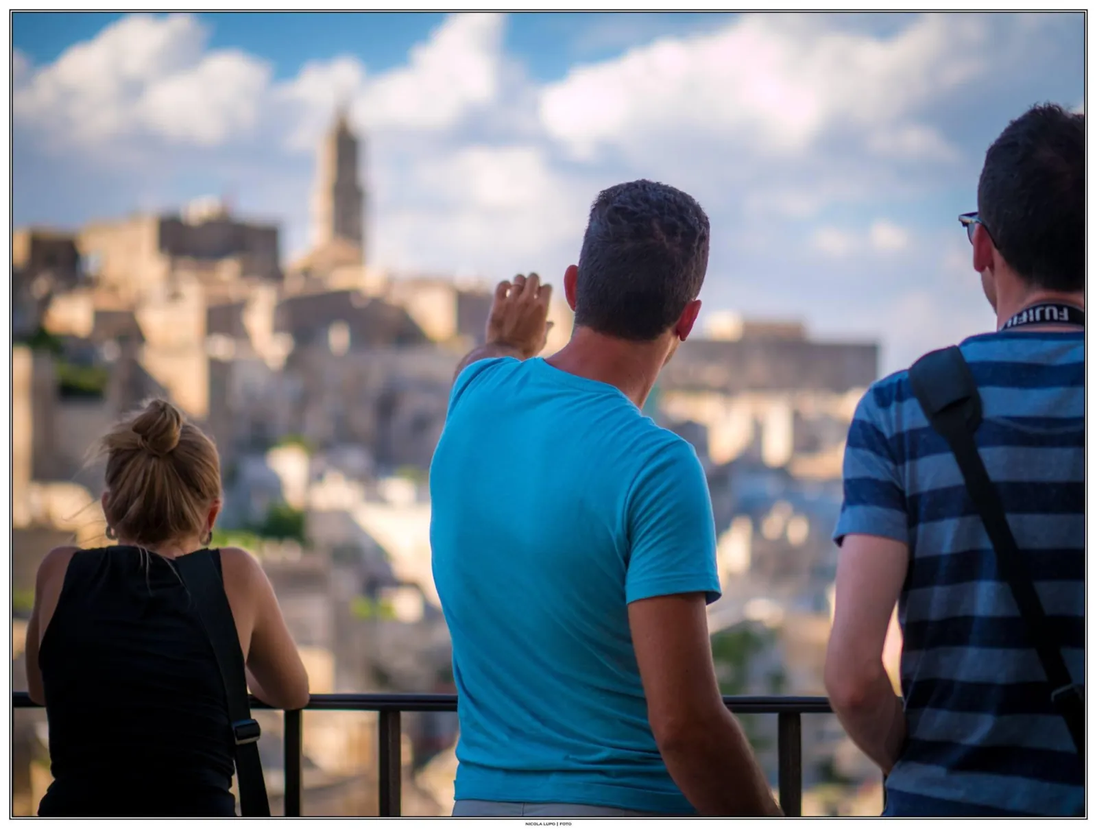 Three visitors lean on a railing looking out over the Sassi rooftops, with a bell tower on the skyline.