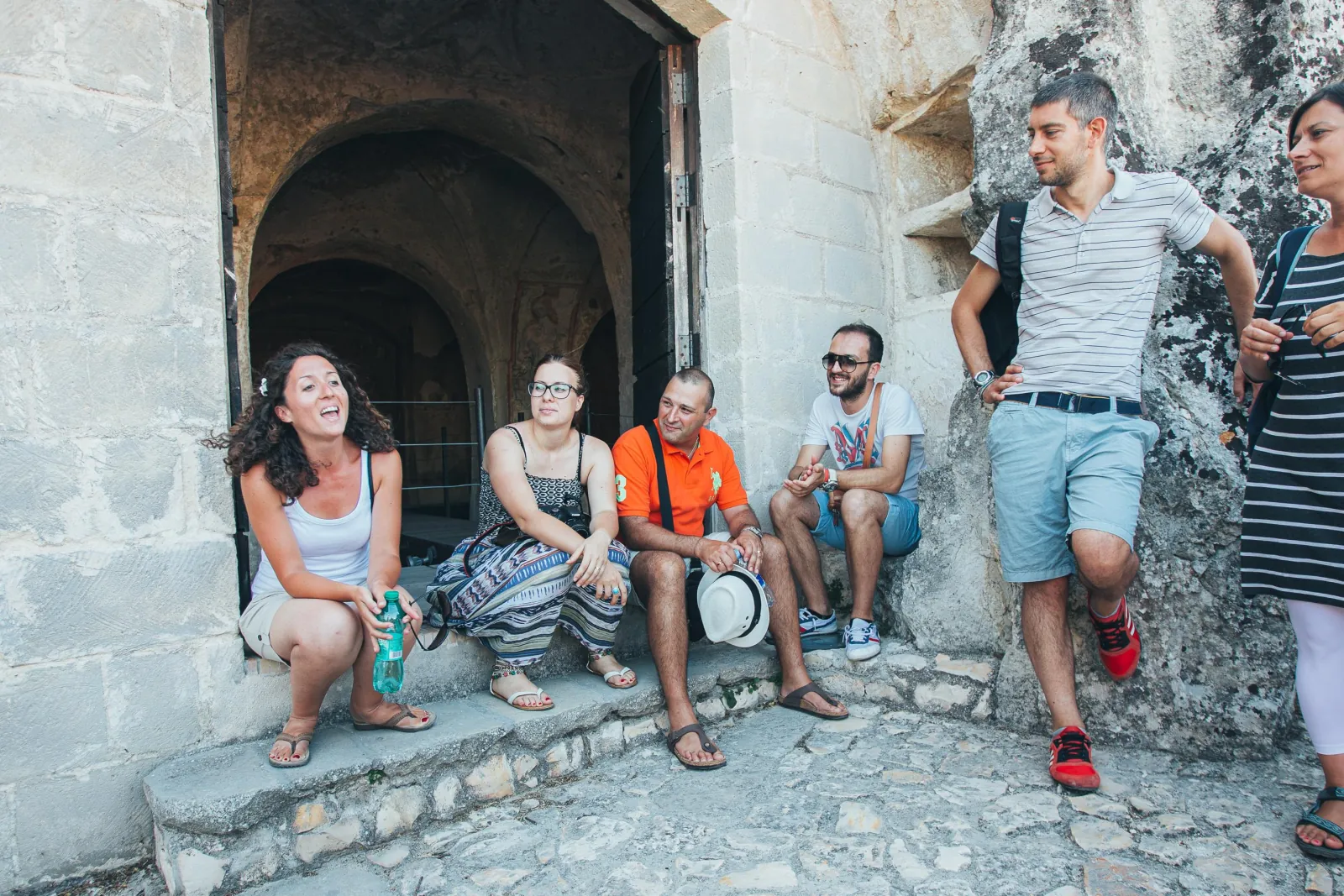 A small group sits and chats on a stone step beside the arched entrance of a cave dwelling in Matera.