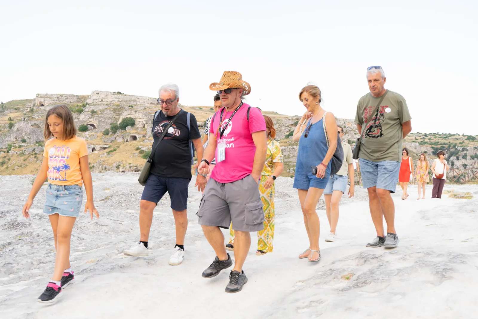 Tour participants walk across a pale limestone plateau with the Murgia hillside and caves behind them near Matera.
