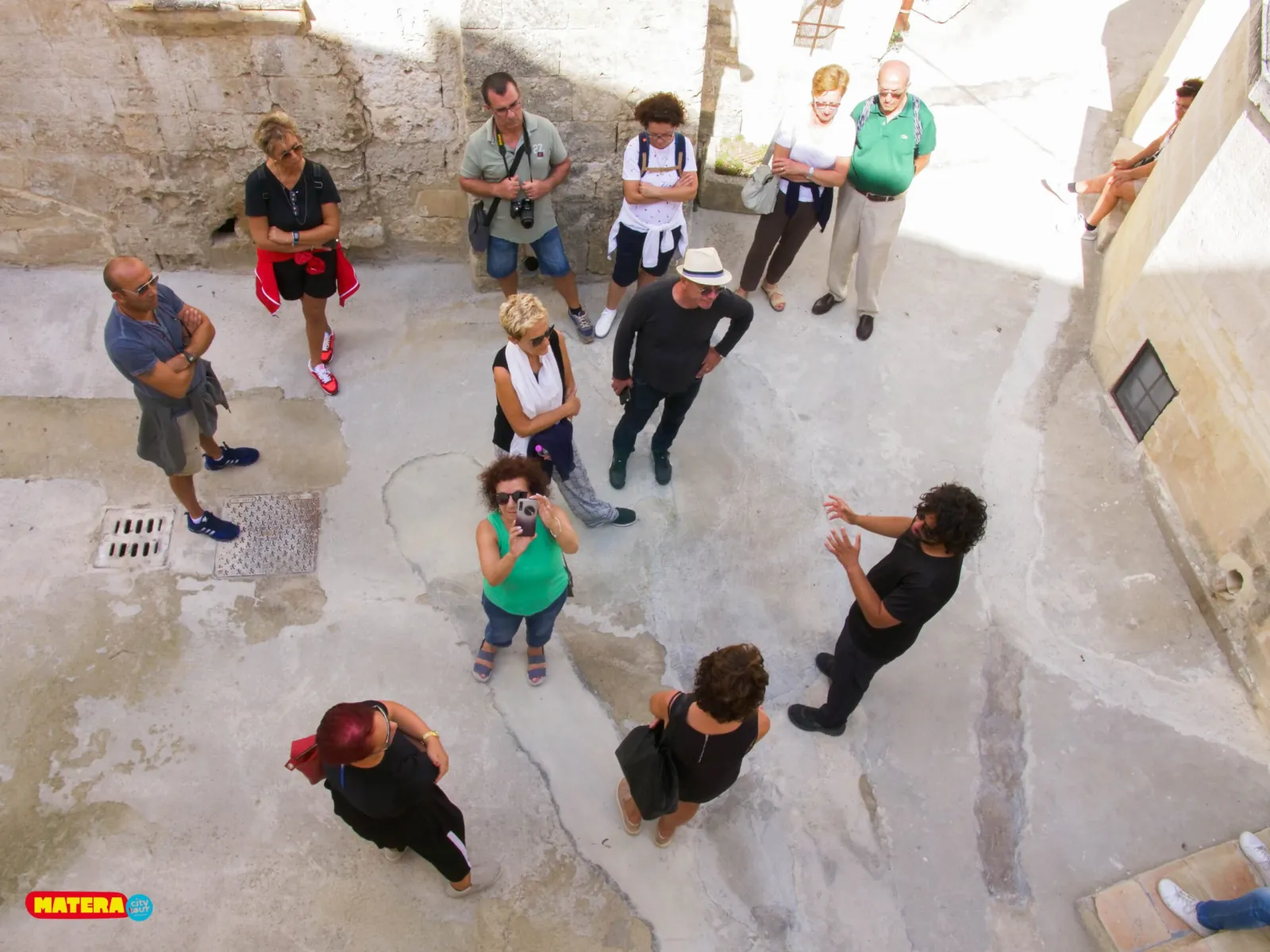 A guide speaks to a group gathered in a whitewashed stone courtyard, seen from above, in the Sassi of Matera.