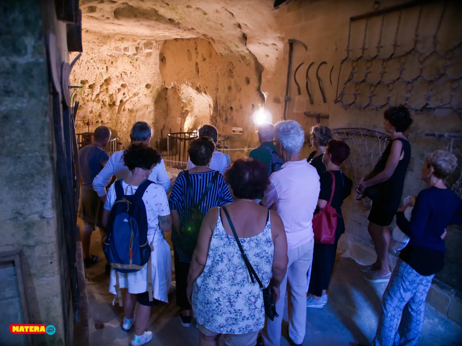 A group stands in a lit rock-cut cave room in Matera with carved niches and old metal tools.