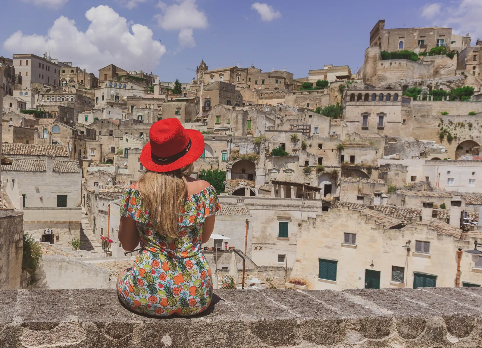 A woman in a red hat and floral dress sits on a wall facing the Sassi's stone houses.