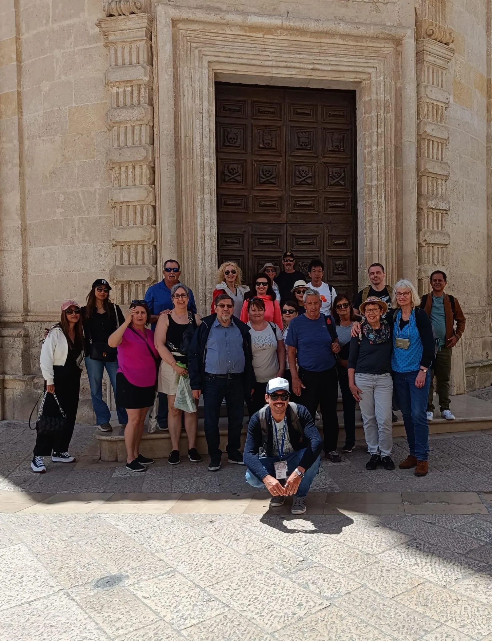 A large tour group poses for a photo in front of a tall carved wooden church door in Matera.