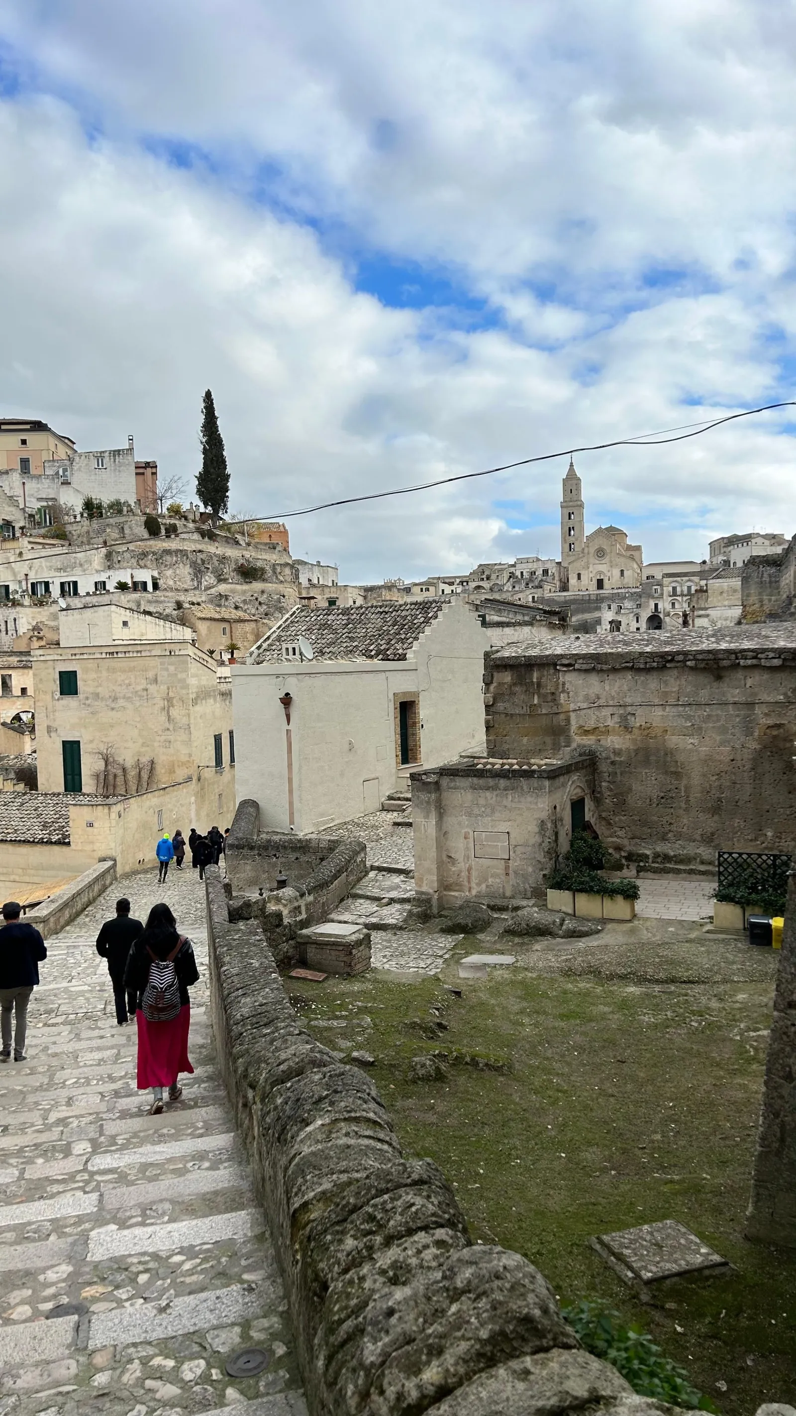 Two visitors walk down a stone stairway between low walls toward the Sassi, with the bell tower in the distance.