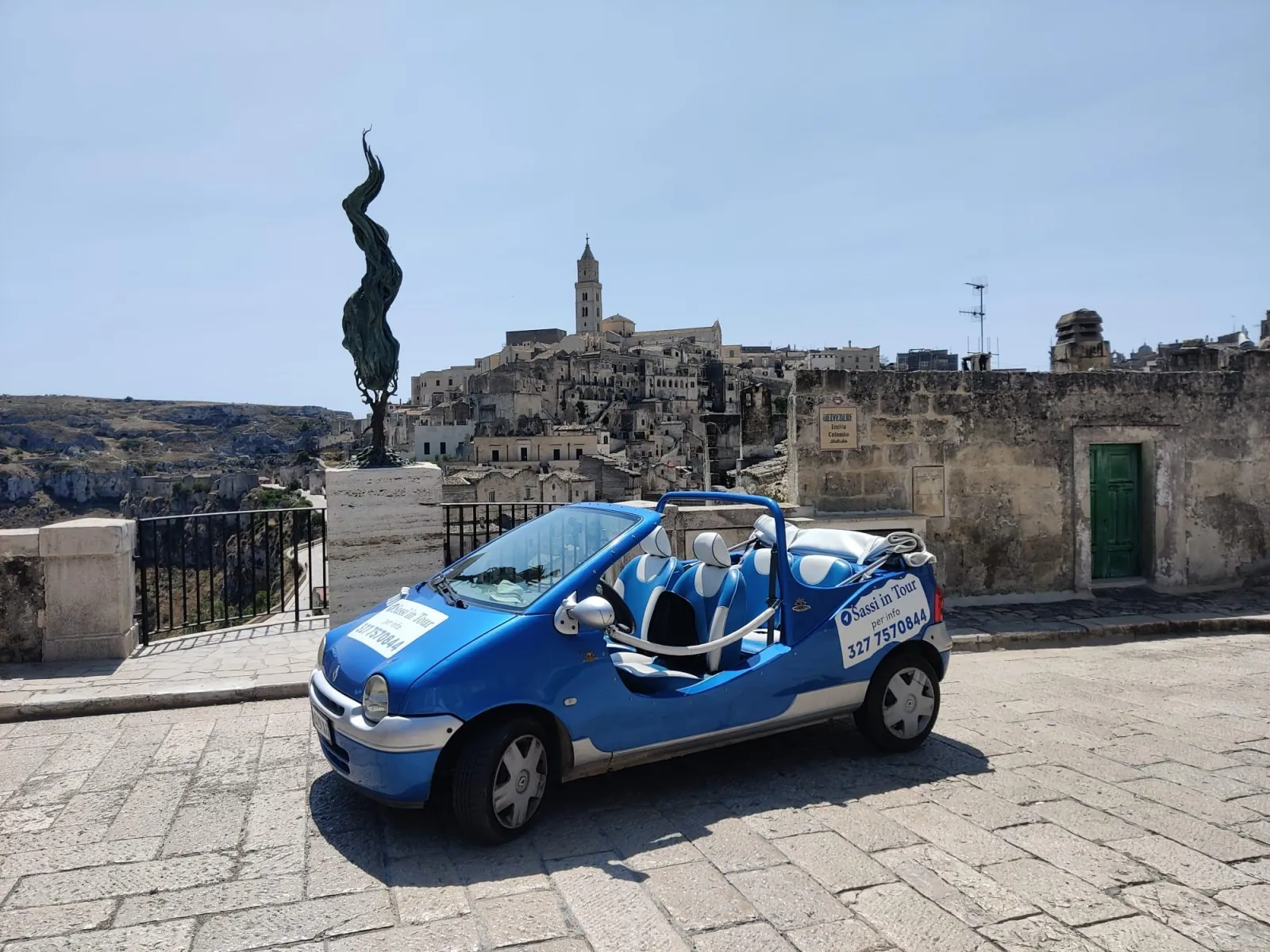 A blue open-top Twingo convertible parked at a Matera viewpoint with a dark bronze sculpture and the old town behind.