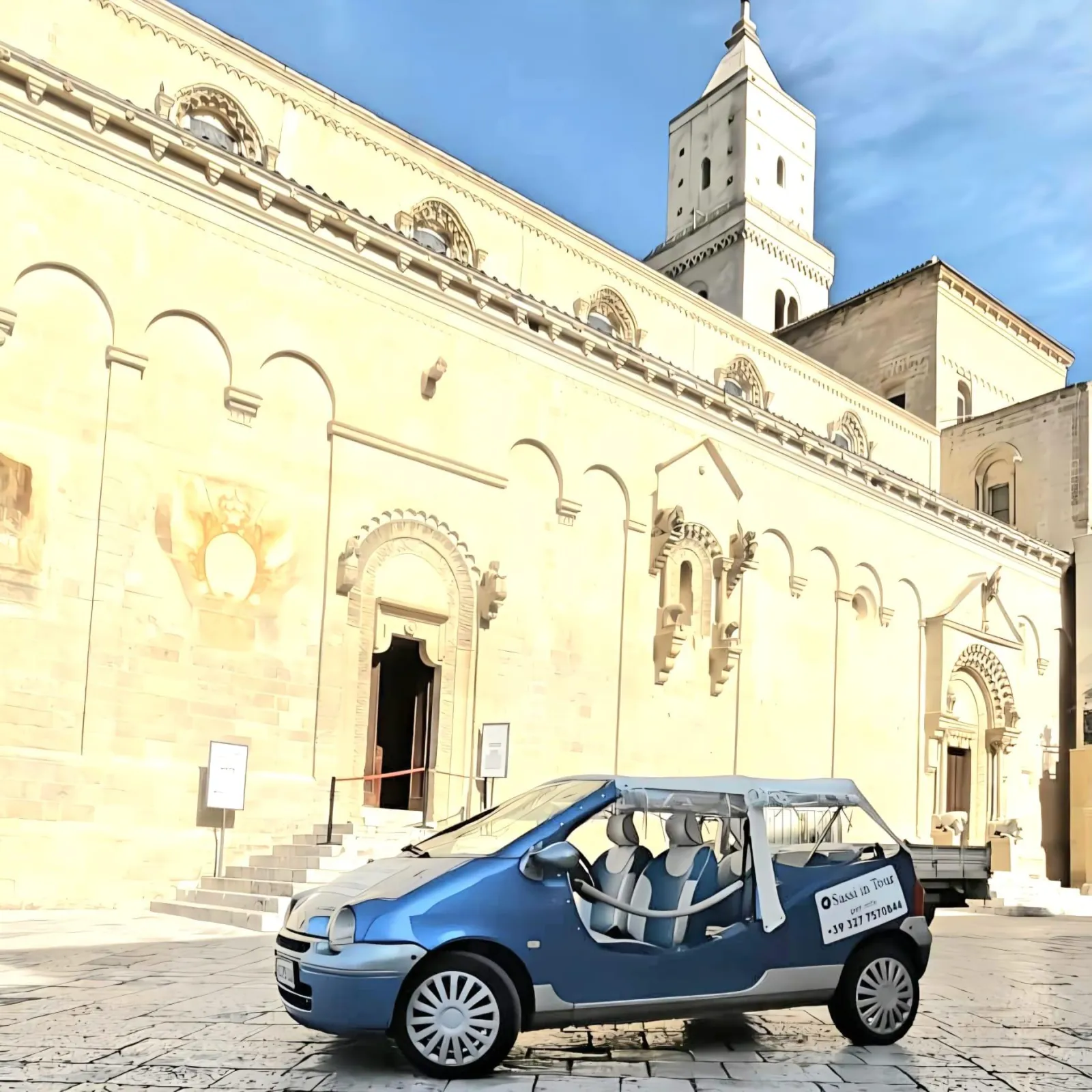 A blue Twingo convertible parked in front of the ornate side facade and bell tower of Matera Cathedral.