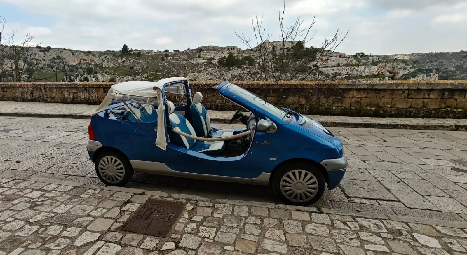 A blue Twingo cabriolet parked on cobblestones beside a stone wall, with the Murgia hillside behind.