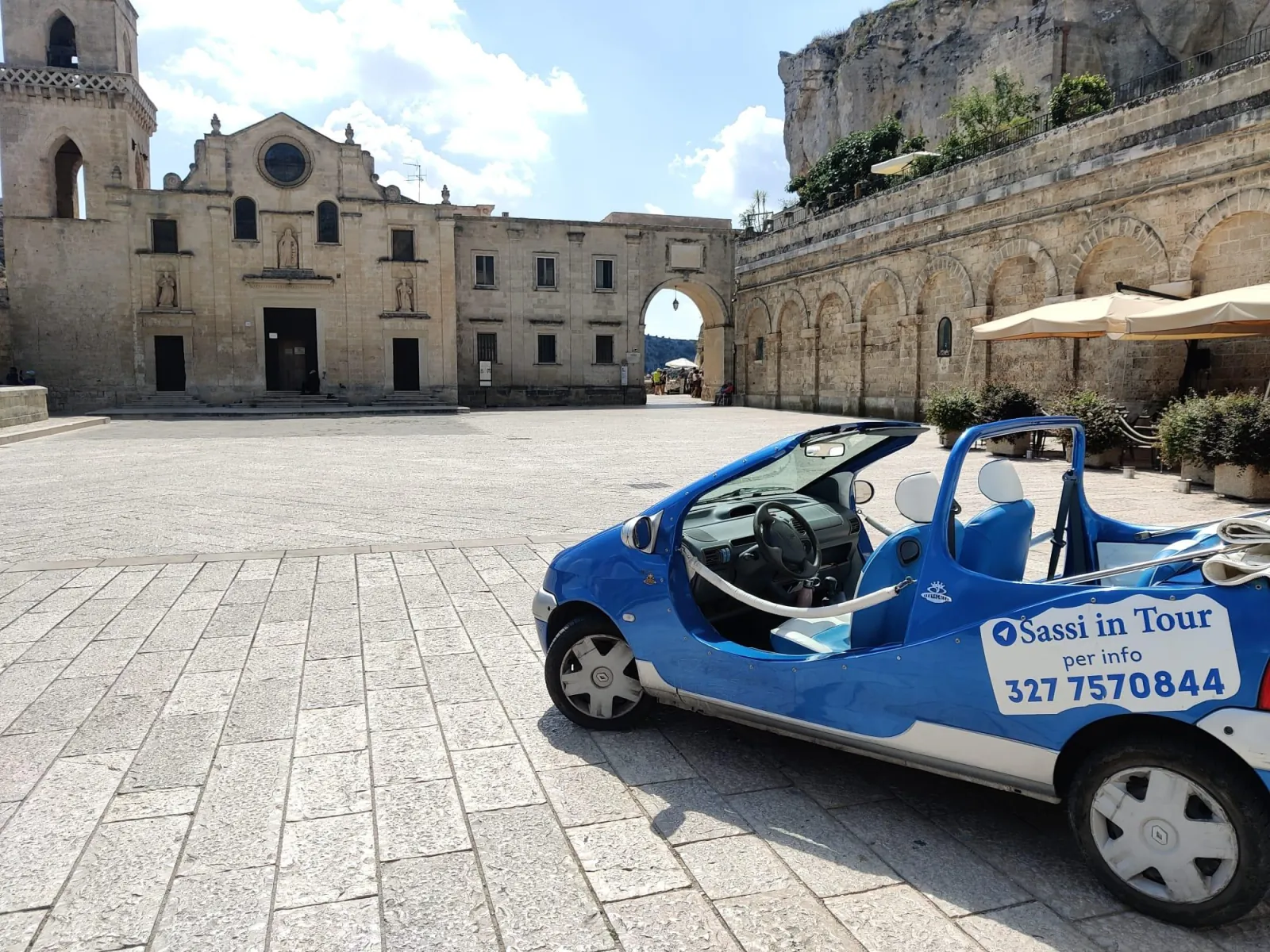 A blue Twingo convertible with a Sassi in Tour door sign in a stone square before a church.
