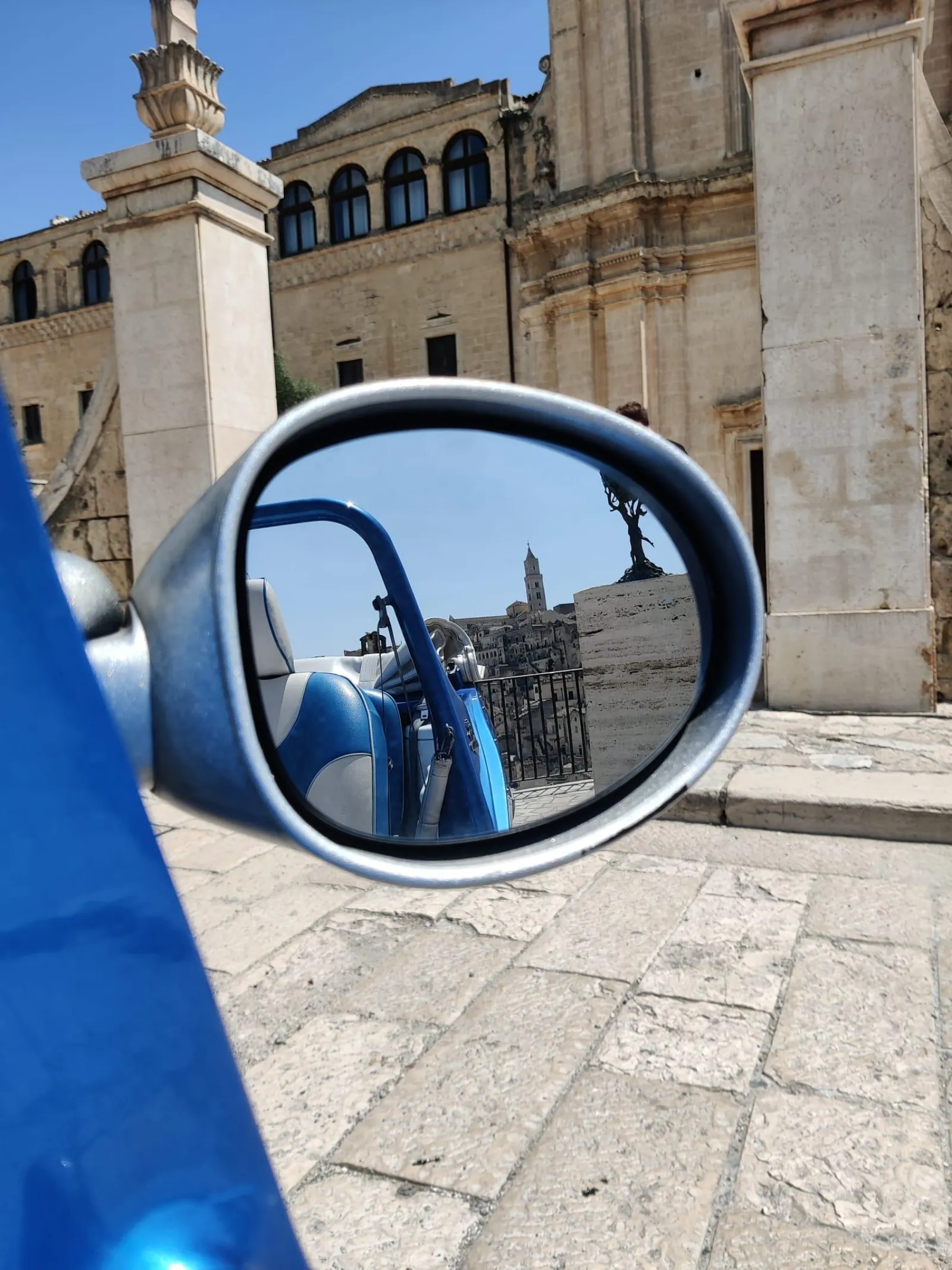 A blue car wing mirror reflects a stone church and tower in a Matera piazza.