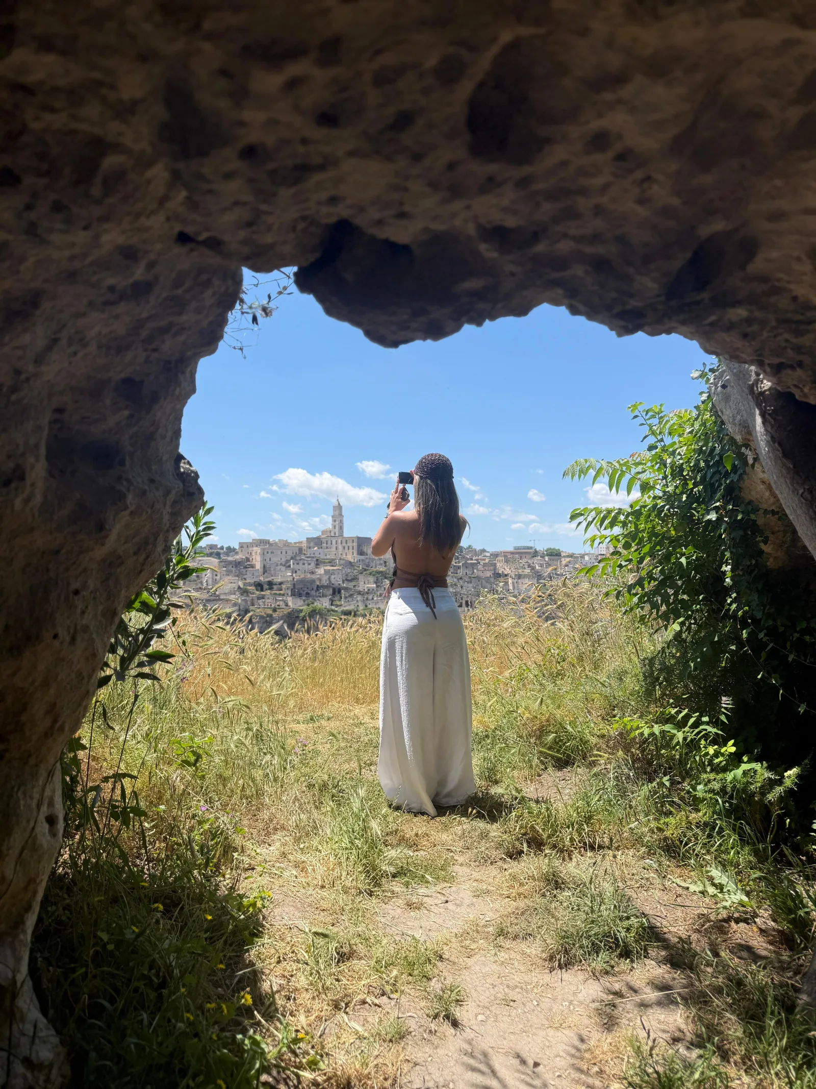 A woman stands inside a rough cave opening looking out at the distant Matera skyline.