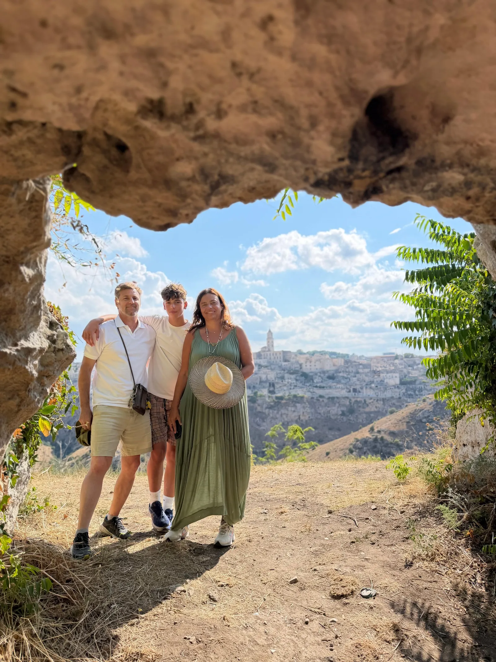 Three visitors pose in a rocky cave opening with a view over the gorge and the distant Sassi.