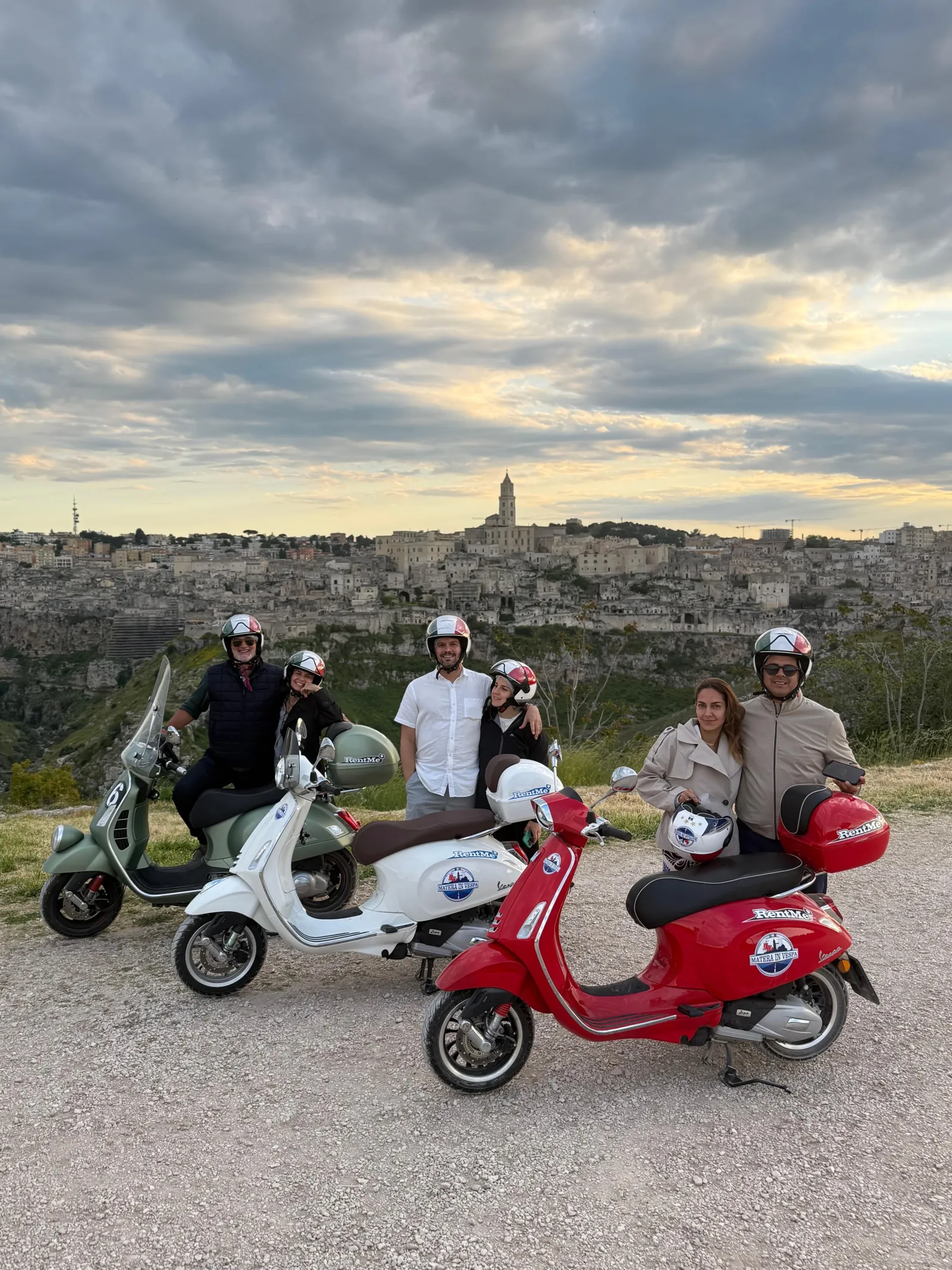 Helmeted riders on white, green and red Vespa scooters stopped on a gravel viewpoint at dusk, with Matera far off.