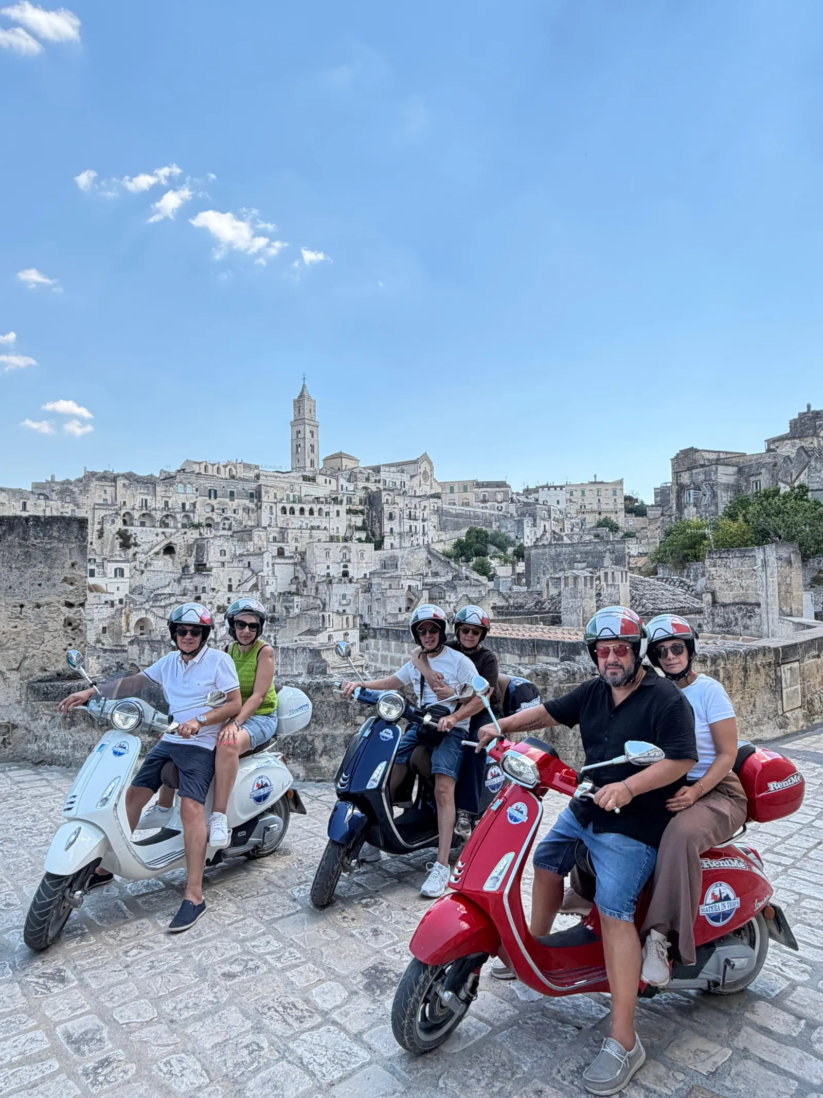 Visitors on white, navy and red Vespa scooters in helmets pose on a stone terrace above the Sassi.